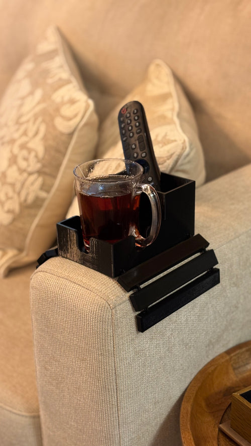 A black plastic armrest organizer tray clipped onto a beige fabric sofa arm, holding a glass mug filled with dark tea and a black remote control, with decorative pillows in the background.