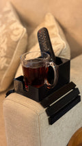 A black plastic armrest organizer tray clipped onto a beige fabric sofa arm, holding a glass mug filled with dark tea and a black remote control, with decorative pillows in the background.
