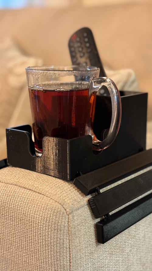 A black plastic armrest organizer tray clipped onto a beige fabric sofa arm, holding a glass mug filled with dark tea and a black remote control, with decorative pillows in the background.